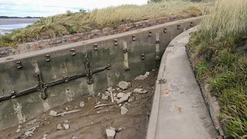 A concrete boat shape sunk in mud, overgrown with grass