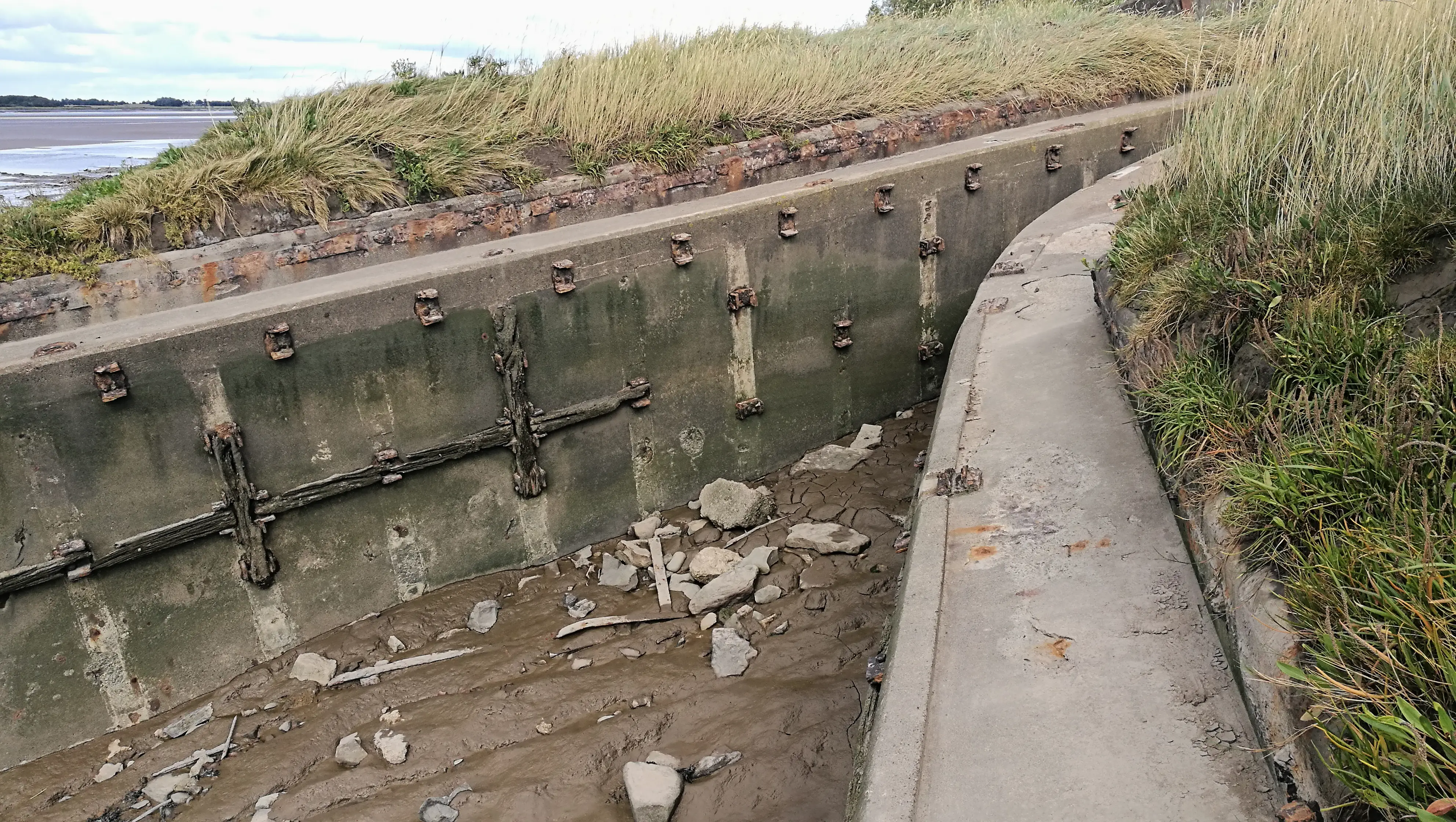 A concrete boat shape sunk in mud, overgrown with grass